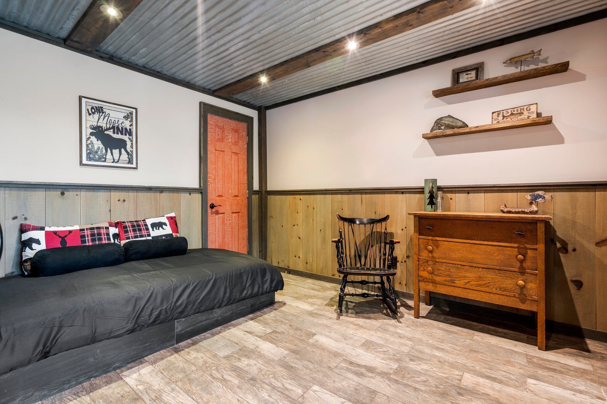 Una habitación con una cama, un aparador de madera, una silla y una pared con un cuadro de un alce y una puerta.