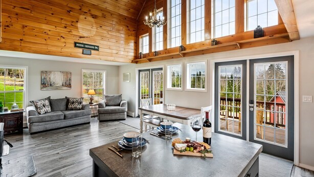 A spacious living area with a high ceiling, large windows, and a dining table set with plates, glasses, and a wine bottle. - S Diamond S Retreat | Huge Backyard & Hot Tub – 2 Miles to Town! (Rangeley)