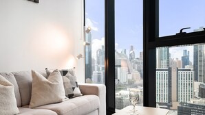 A modern living room with a sofa, pillows, a glass of wine, and a view of a cityscape through large windows. - Stylish Southbank 1-bed Flat With Luxury Amenities (Melbourne)