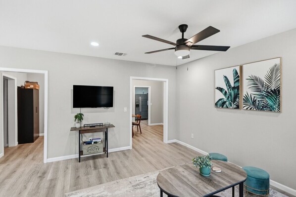 A modern living room with a flat-screen TV, a ceiling fan, and a dining area with a wooden table and blue chairs. - Spacious 4BR Haven Perfect for Groups Near DT (San Antonio)