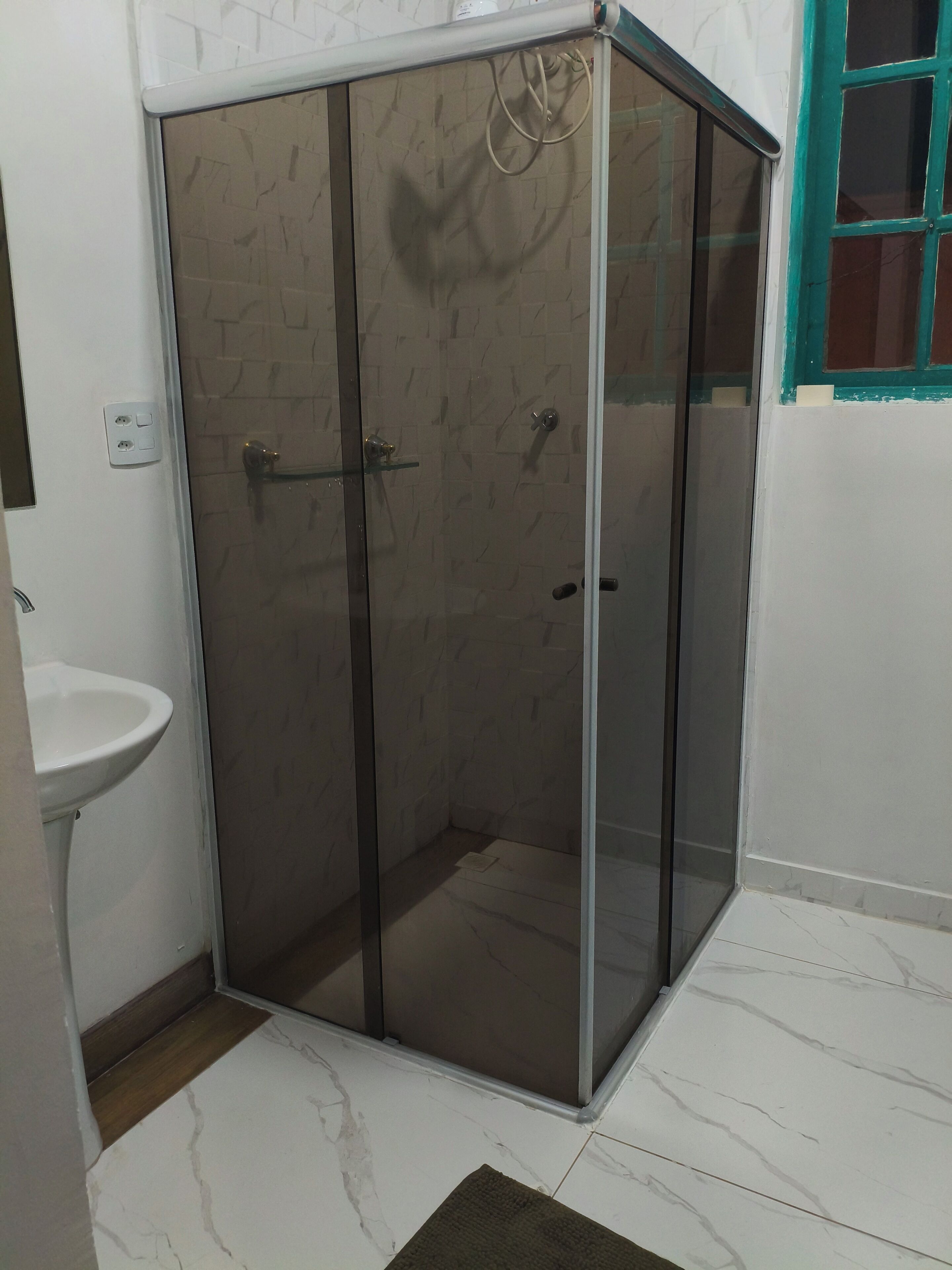 Een badkamer met een douche met glazen wand, een wit wastafeltje en een betegelde vloer.
