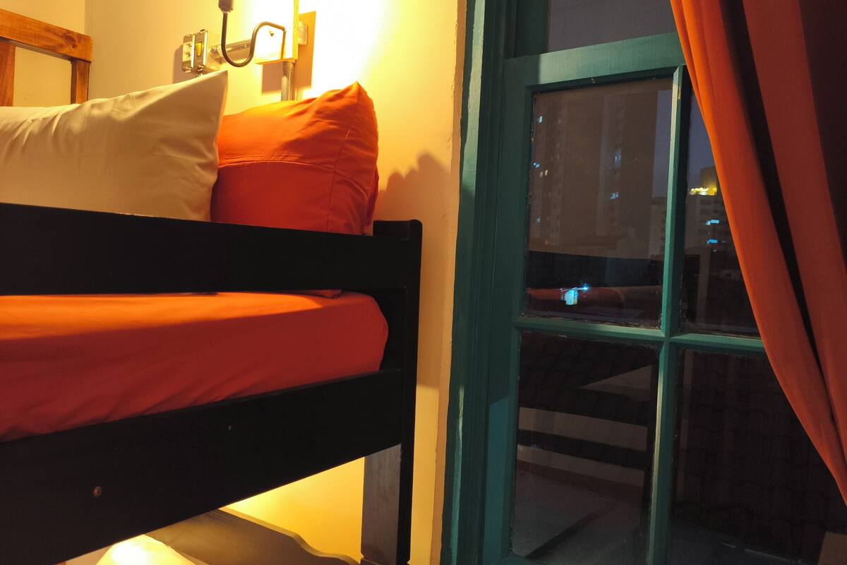 Un letto a castello con materasso e cuscini rossi, una lampada da comodino e una finestra con tende.