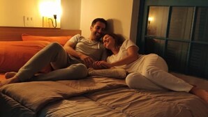 Una pareja recostada en una cama en una habitación de hotel, con un ventanal grande y una lámpara fijada a la pared.