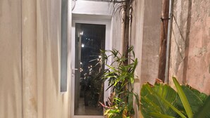 Un camí de maó estret que condueix a una porta, flanquejat per plantes en testos i una paret amb canonades a la vista.
