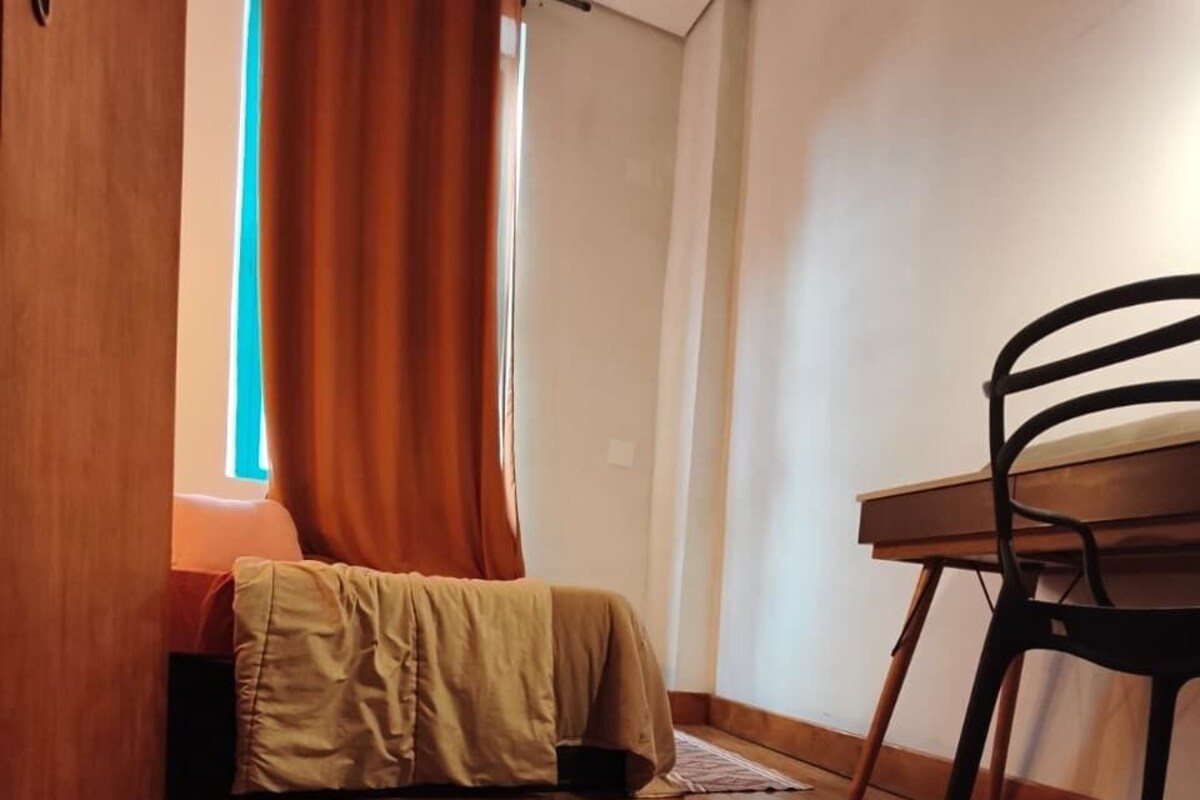 Una habitación con piso de madera, una cama con manta color beige, una silla de madera y una ventana con cortinas naranjas.