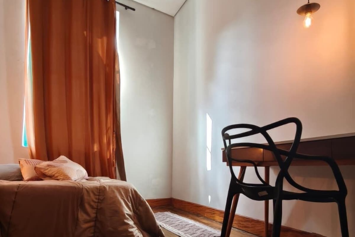 Una habitación con cama, una silla, piso de madera, ventana con cortinas y una lámpara colgante.