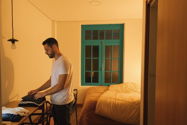 A person folding clothes at a table in a room with a bed, a window with green shutters, and a hanging light fixture.