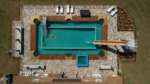 An aerial view of a rectangular swimming pool with a diving board, surrounded by a tiled deck and lounge chairs.