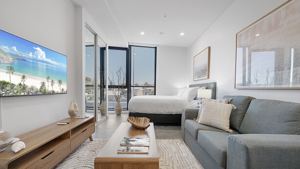 Une chambre à coucher moderne avec un lit, un canapé gris, une table basse en bois et un grand téléviseur fixé au mur.