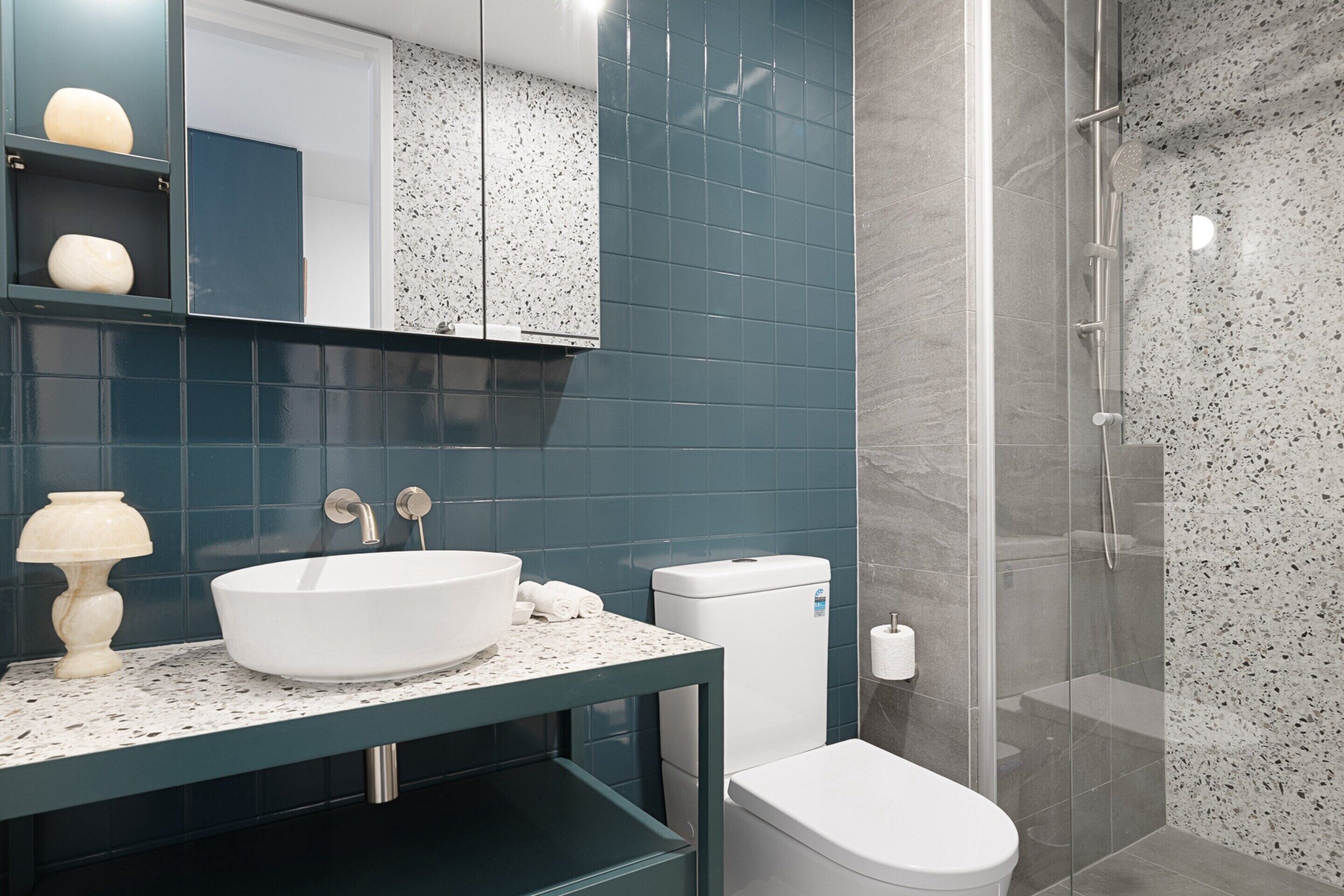 A modern bathroom with a white sink, dark blue tiles, a glass shower enclosure, and a marble backsplash.