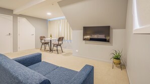 A modern living room with a blue sofa, a dining area with a table and chairs, and a TV mounted on the wall. - Woolshed Suites (Lake Tekapo)