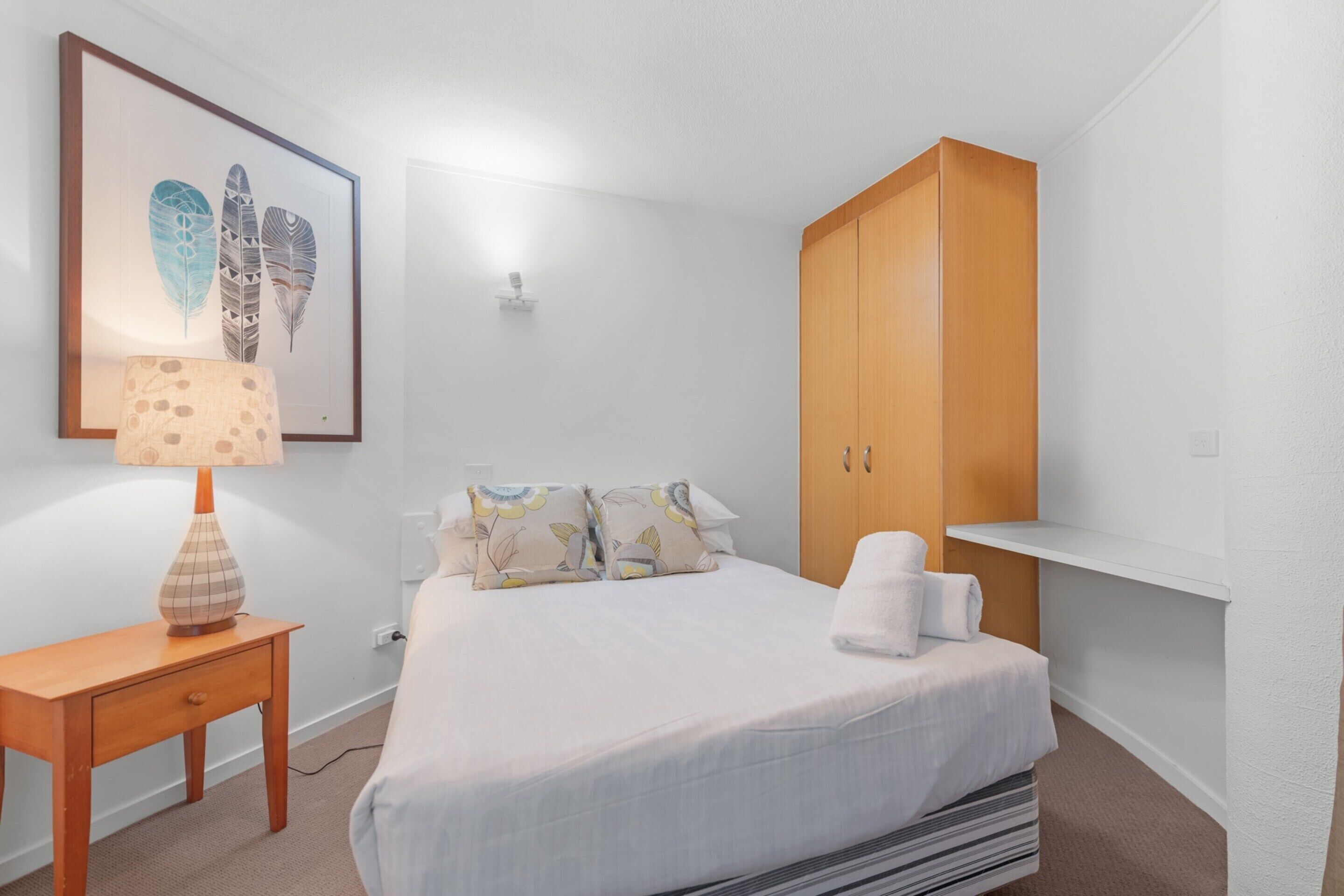 A bedroom with a bed, a wooden wardrobe, a nightstand with a lamp, and a framed artwork on the wall. - Stylish 3BR Oceanfront Stay by Mooloolaba Esp (Sunshine Coast)