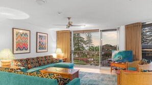 A spacious living room with a teal sofa, a coffee table, and a television on a stand. There is a balcony with a view of trees and a cityscape. - Stylish 3BR Oceanfront Stay by Mooloolaba Esp (Sunshine Coast)