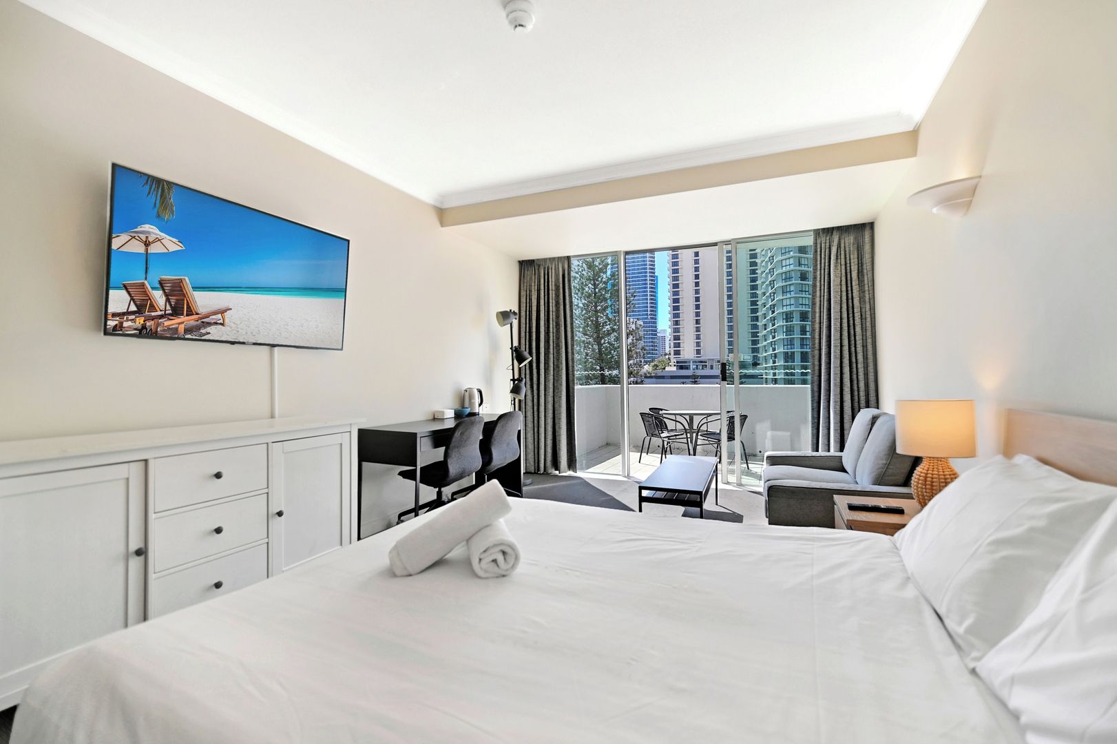 A hotel room with a bed, a desk, a chair, a small table, a lamp, and a painting of a beach scene.