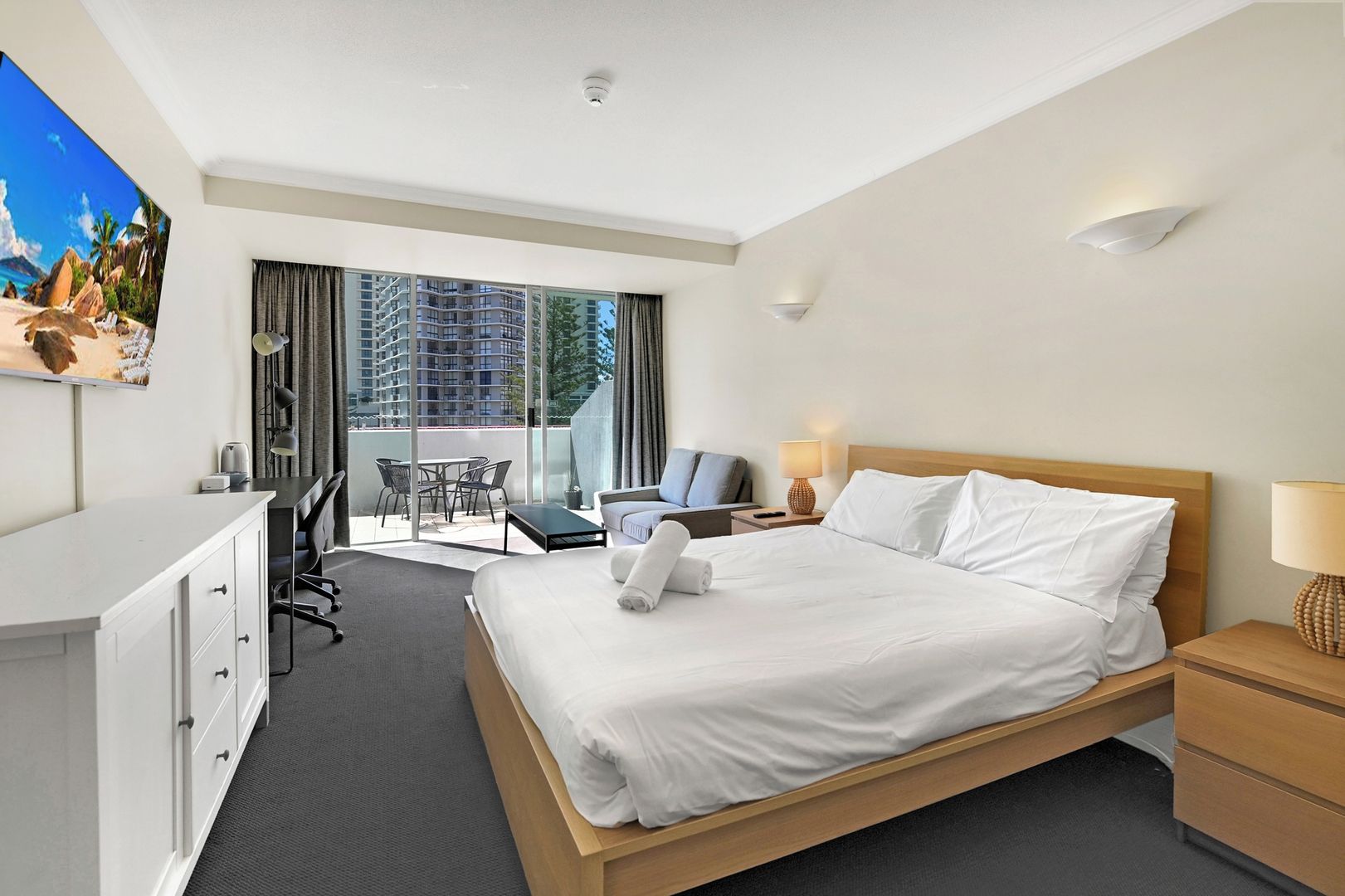 A hotel room with a bed, desk, chair, and a view of buildings through large windows.