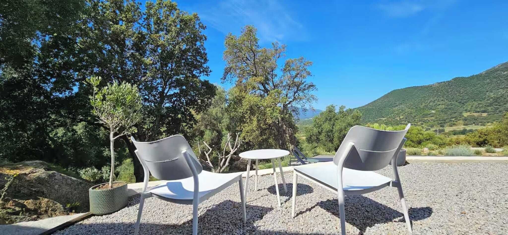 Two white chairs and a small round table on a gravel surface, surrounded by trees and a view of a hilly landscape.