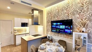 Una cocina moderna con televisor integrado mostrando servicios de streaming, un comedor con mesa lista para usar, y un papel tapiz estampado.