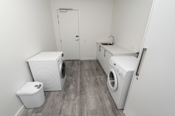 A compact laundry room with two washing machines, a sink, and a trash bin.