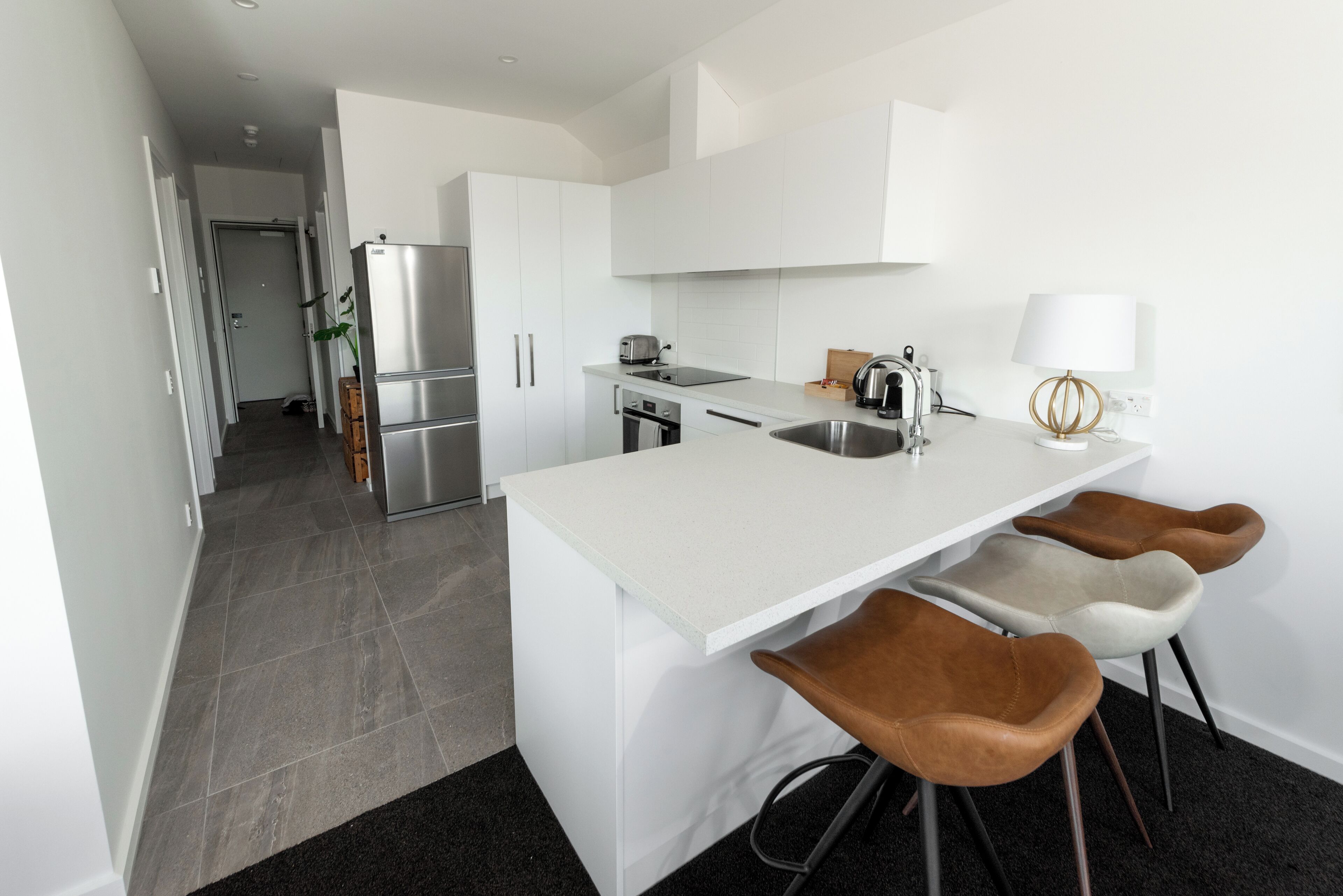 A modern kitchen with white cabinetry, a stainless steel refrigerator, a built-in oven, and a sink. There are three stools with brown seats and a white countertop.