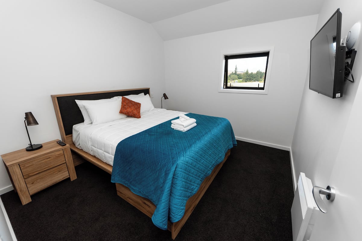 A neatly made bed with a blue blanket, a wooden nightstand with a lamp, and a small window with a view of greenery.