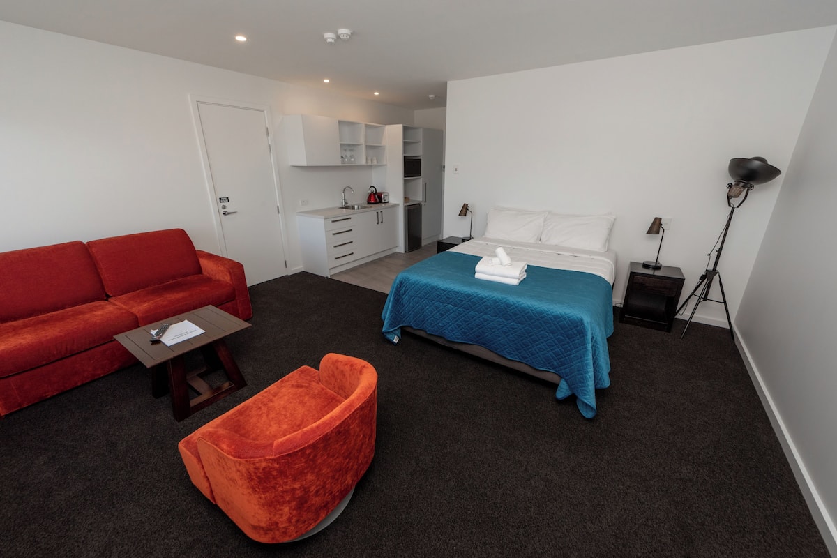 A modern hotel room with a red sofa, a blue bed, a small orange chair, a kitchenette, and a desk with a lamp.