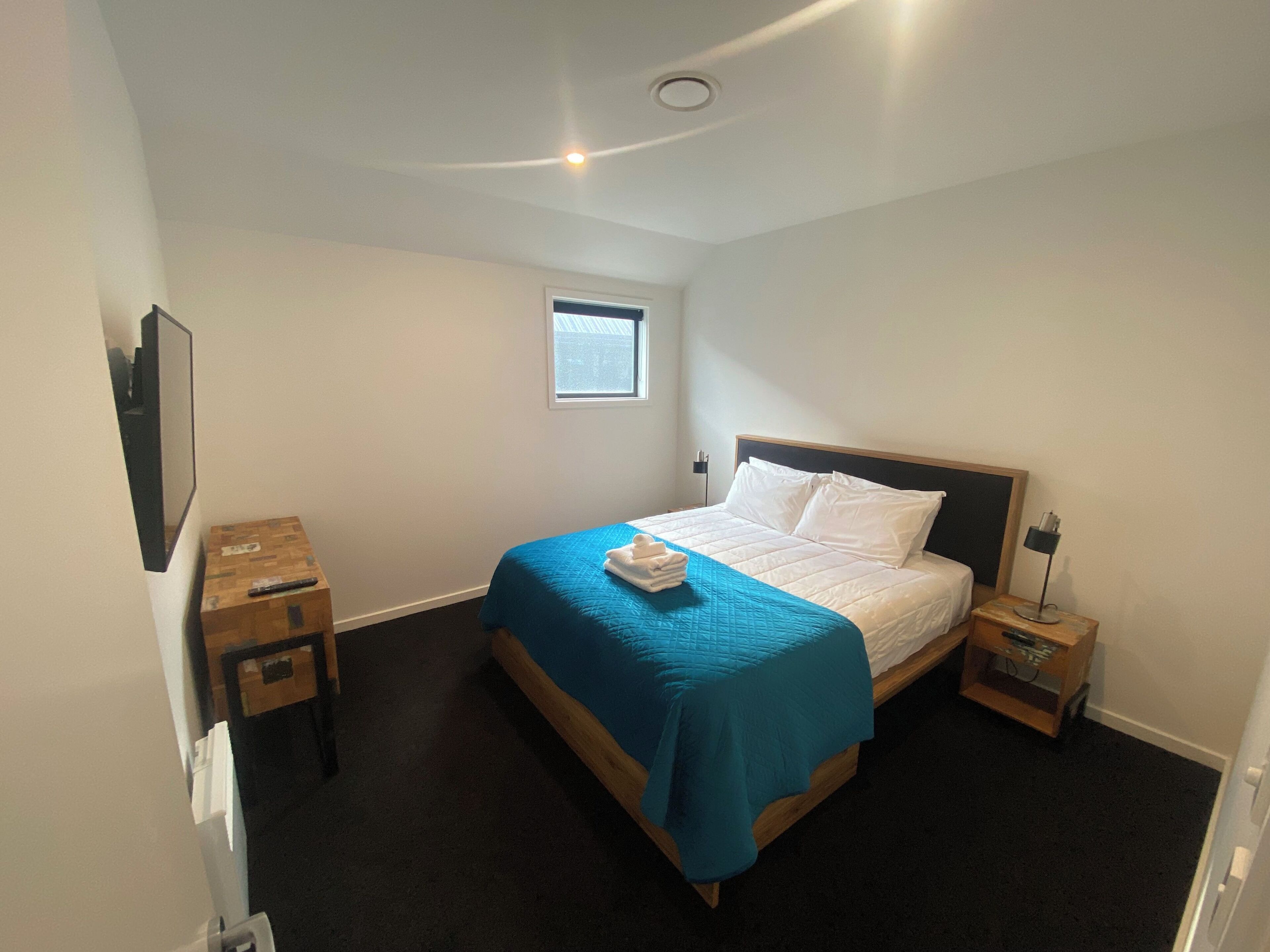 Un dormitorio con cama, una mesita de noche pequeña, una ventana y un televisor instalado en la pared.