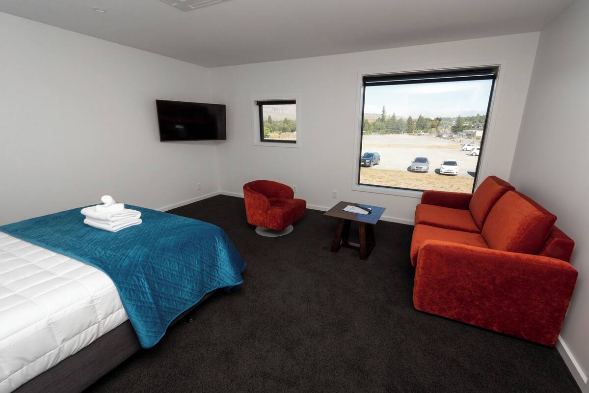 A hotel room with a bed, a red armchair, a sofa, a small table, and a TV on the wall.