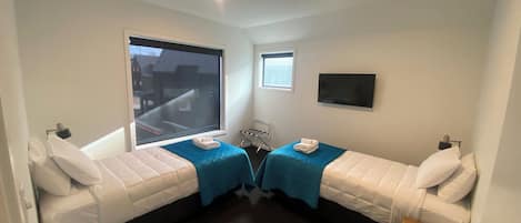 Habitación pequeña y funcional con dos camas individuales, una mesita, una silla, un televisor montado en la pared y una ventana con vistas a la calle.