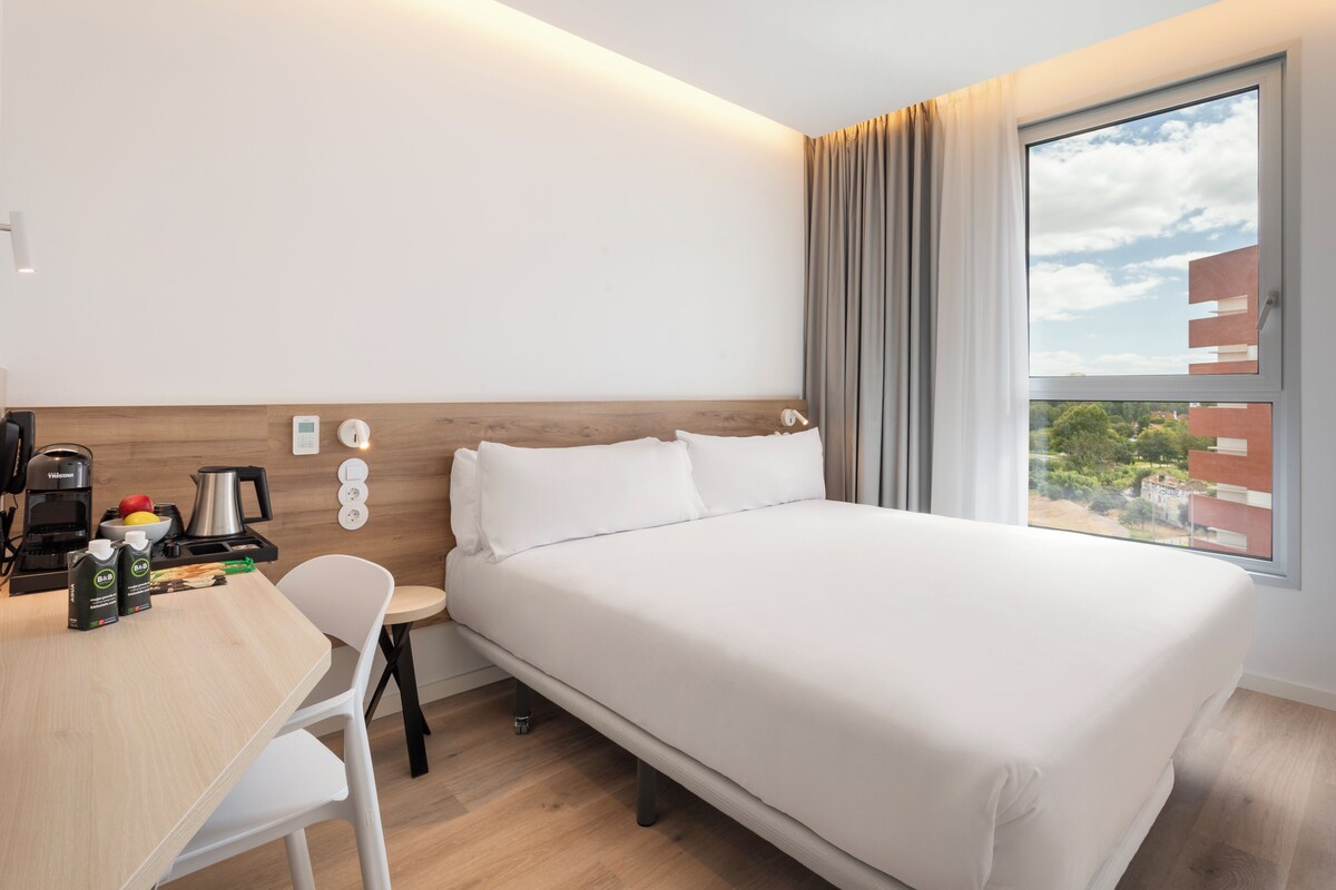 A hotel room with a bed, a desk with a coffee maker, a kettle, and a fruit bowl.