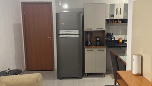 Uma cozinha compacta com geladeira, cafeteira e micro-ondas. Há uma porta de madeira e uma mesa pequena com uma cadeira.