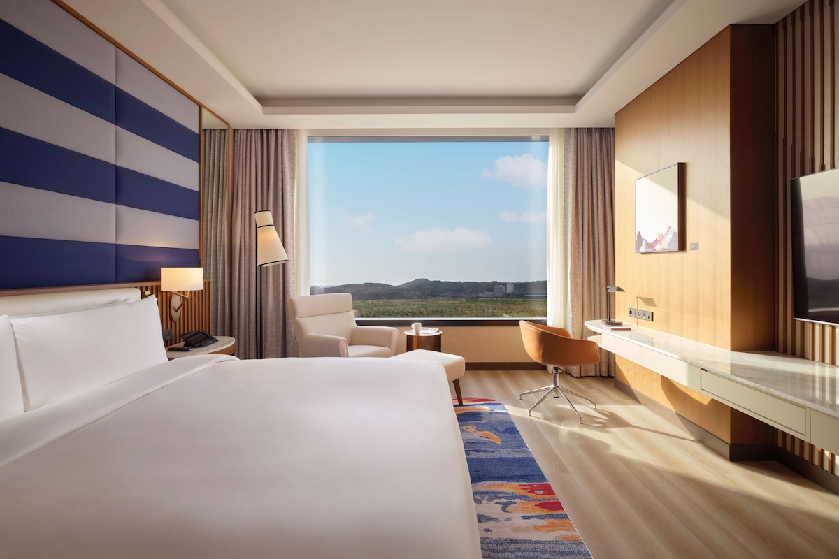 A hotel room with a large bed, a desk, a chair, and a view of the outdoors.