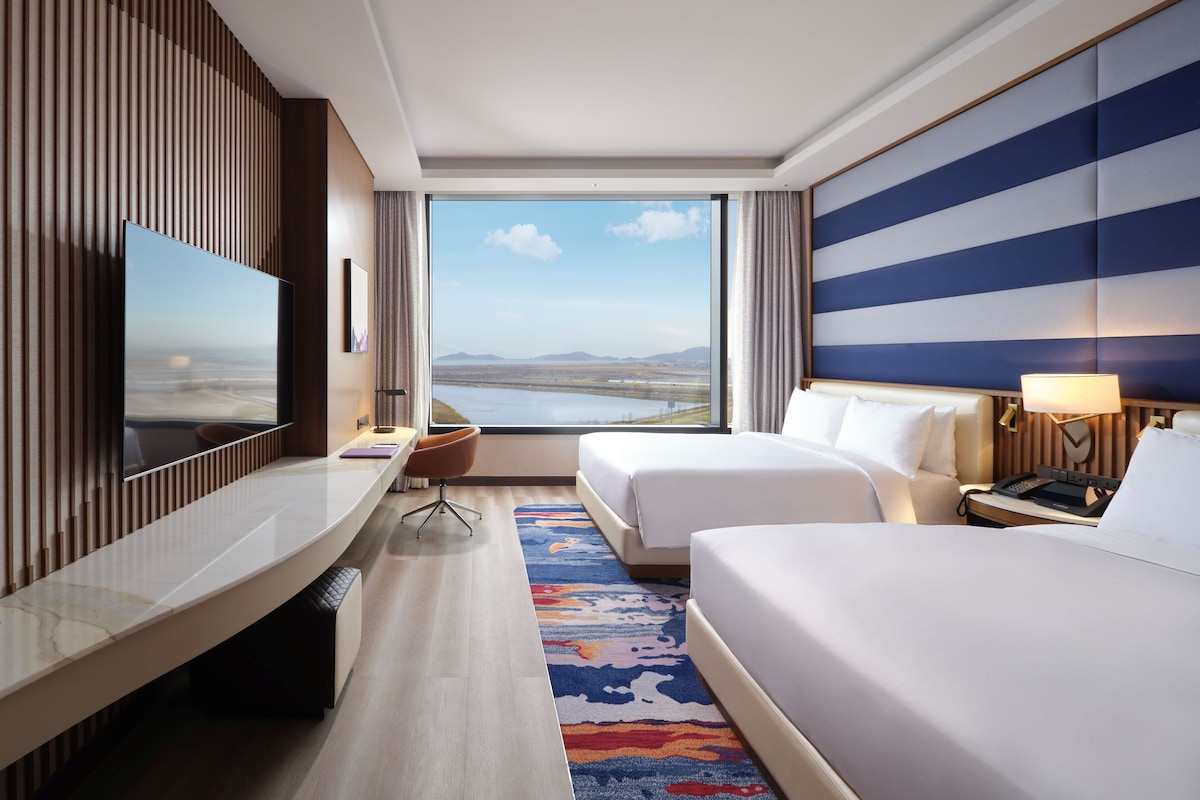 A hotel room with a large window, a flat-screen TV, two beds, a desk, and a chair.