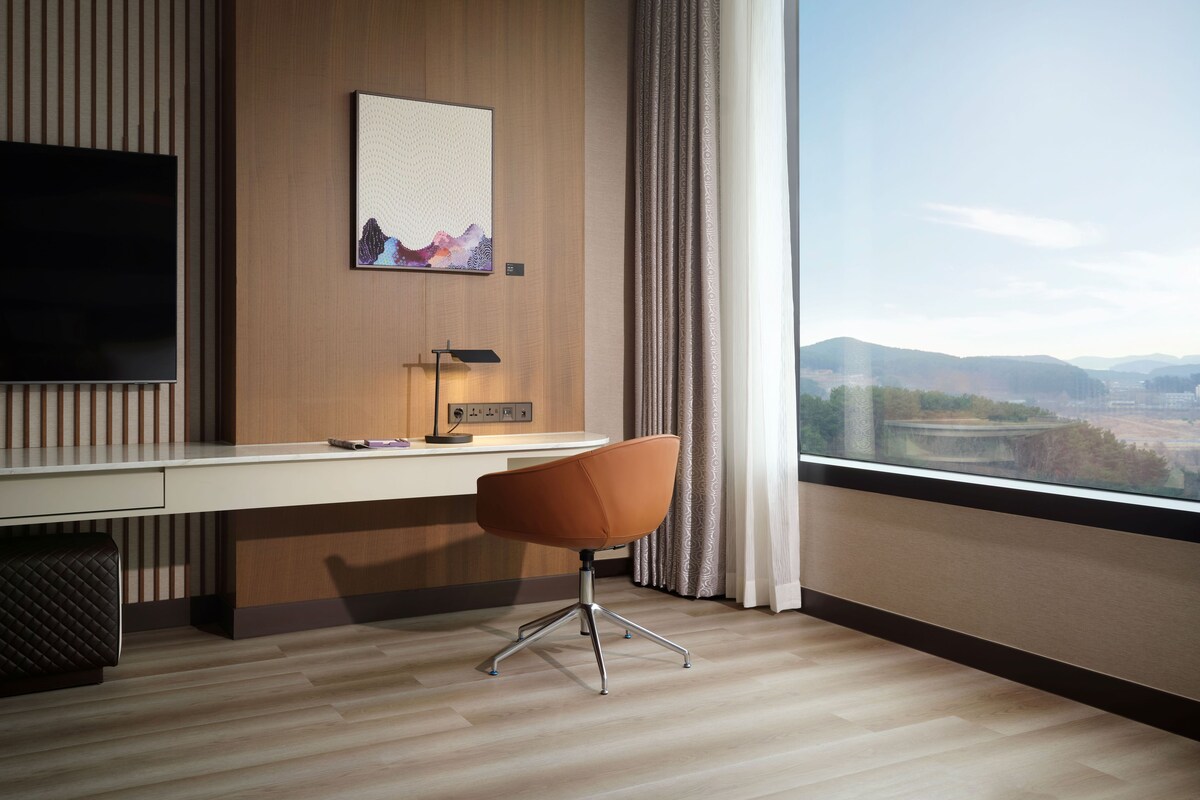 A modern hotel room with a large desk, a flat-screen TV, a desk lamp, and a view of a cityscape through large windows.