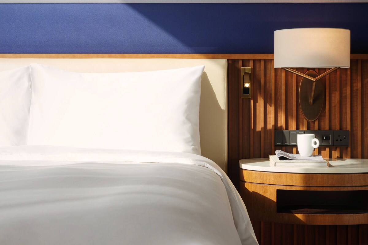 A hotel room with a neatly made bed, a bedside table with a lamp, and a wall-mounted clock.