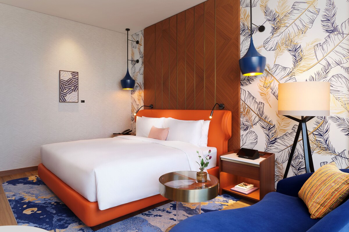 A modern hotel room with a large bed, a bedside table, a nightstand, a floor lamp, and a sofa.
