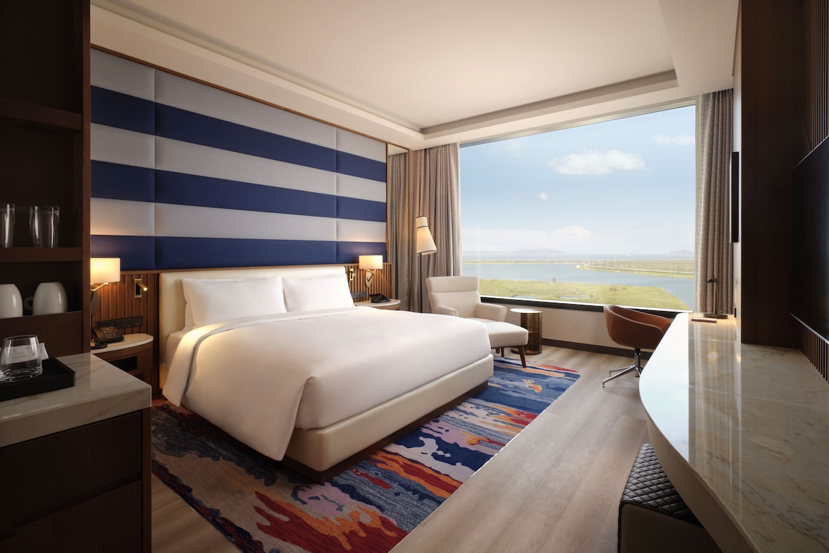A modern hotel room with a large bed, a desk, a chair, a TV, and a view of the landscape.