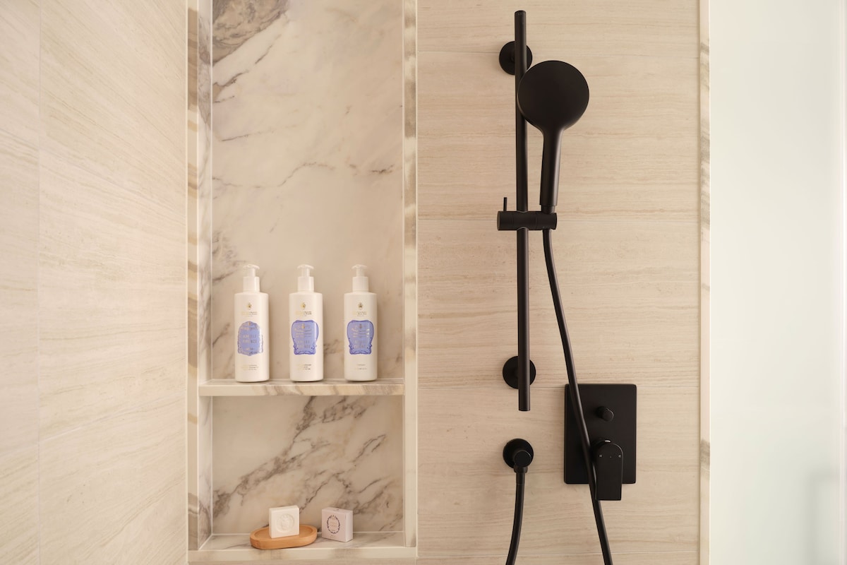 A bathroom with a marble wall, a shelf with toiletries, and a black showerhead.