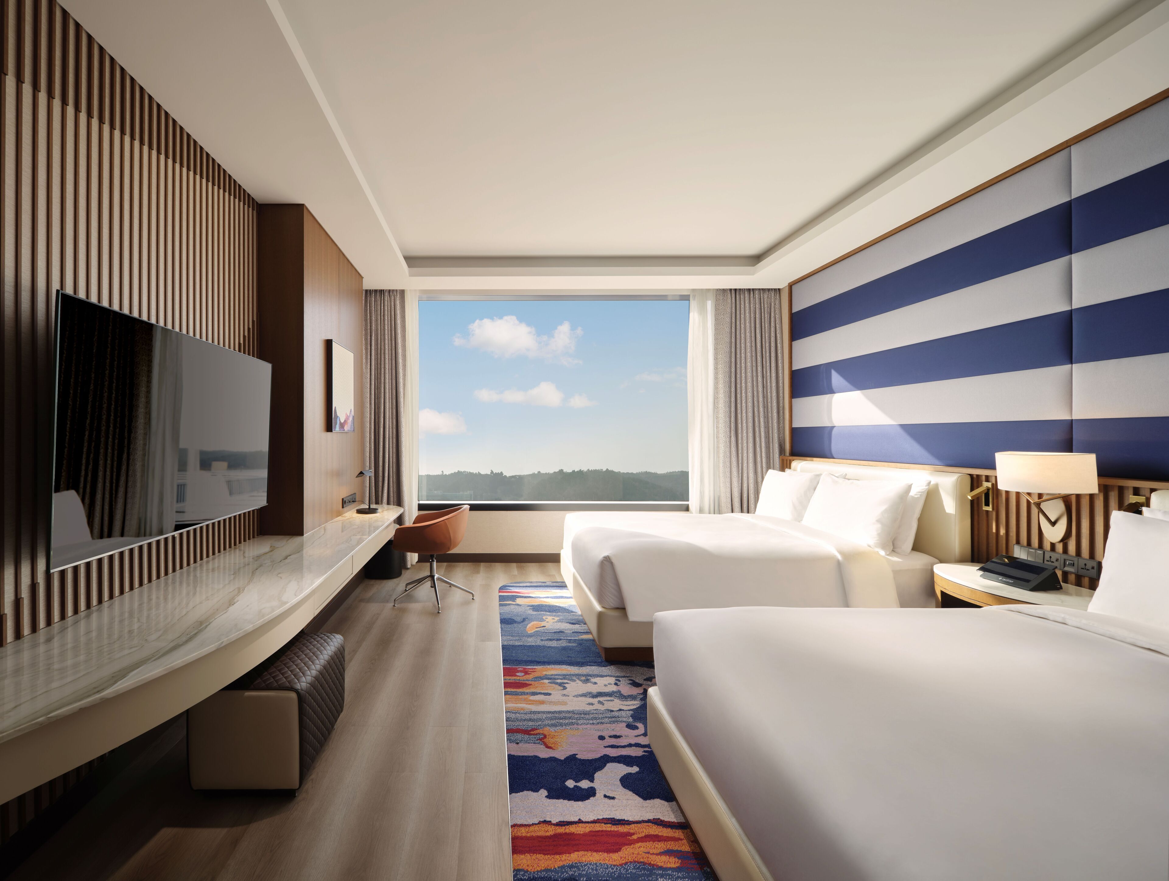 A hotel room with a large TV, two beds, a desk, and a view of the outdoors.