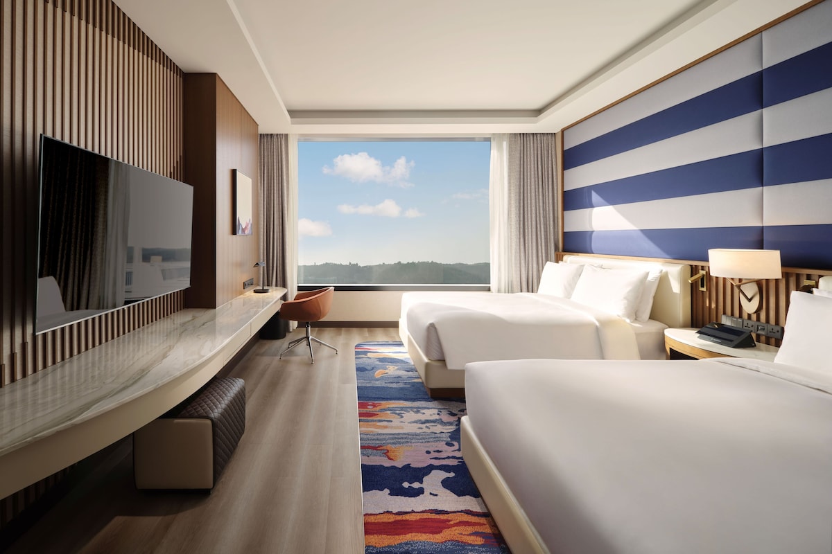 A hotel room with a large TV, two beds, a desk, and a view of the outdoors.