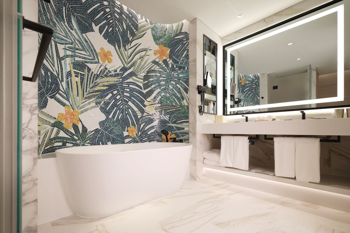 A modern bathroom with a freestanding bathtub, a large mirror, and a decorative leaf-patterned wall.