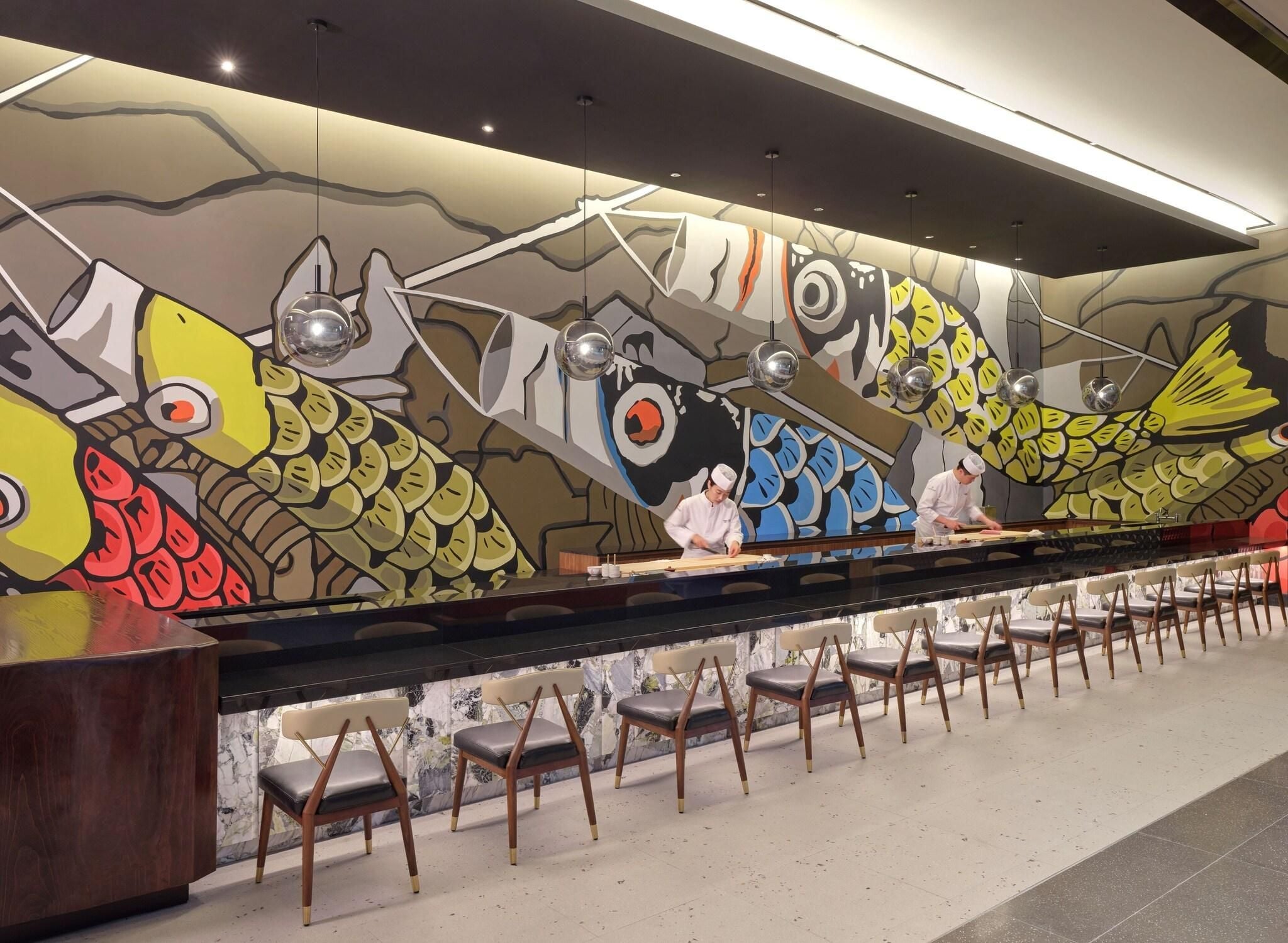 Interior de restaurant amb un comptador llarg, diverses cadires i un mural de peixos de colors a la paret.