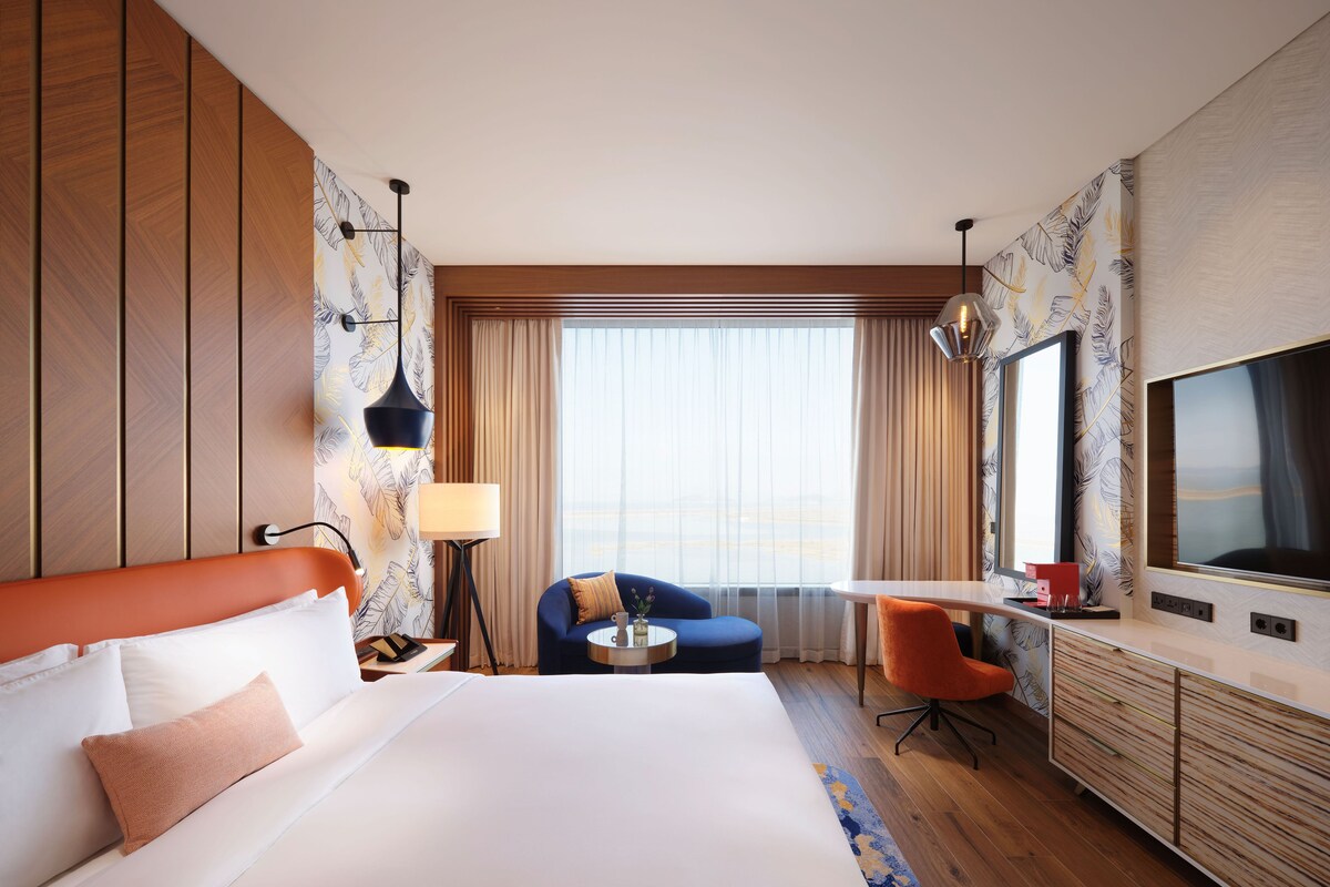 A hotel room with a large bed, a desk, a chair, a TV, and a window with curtains.
