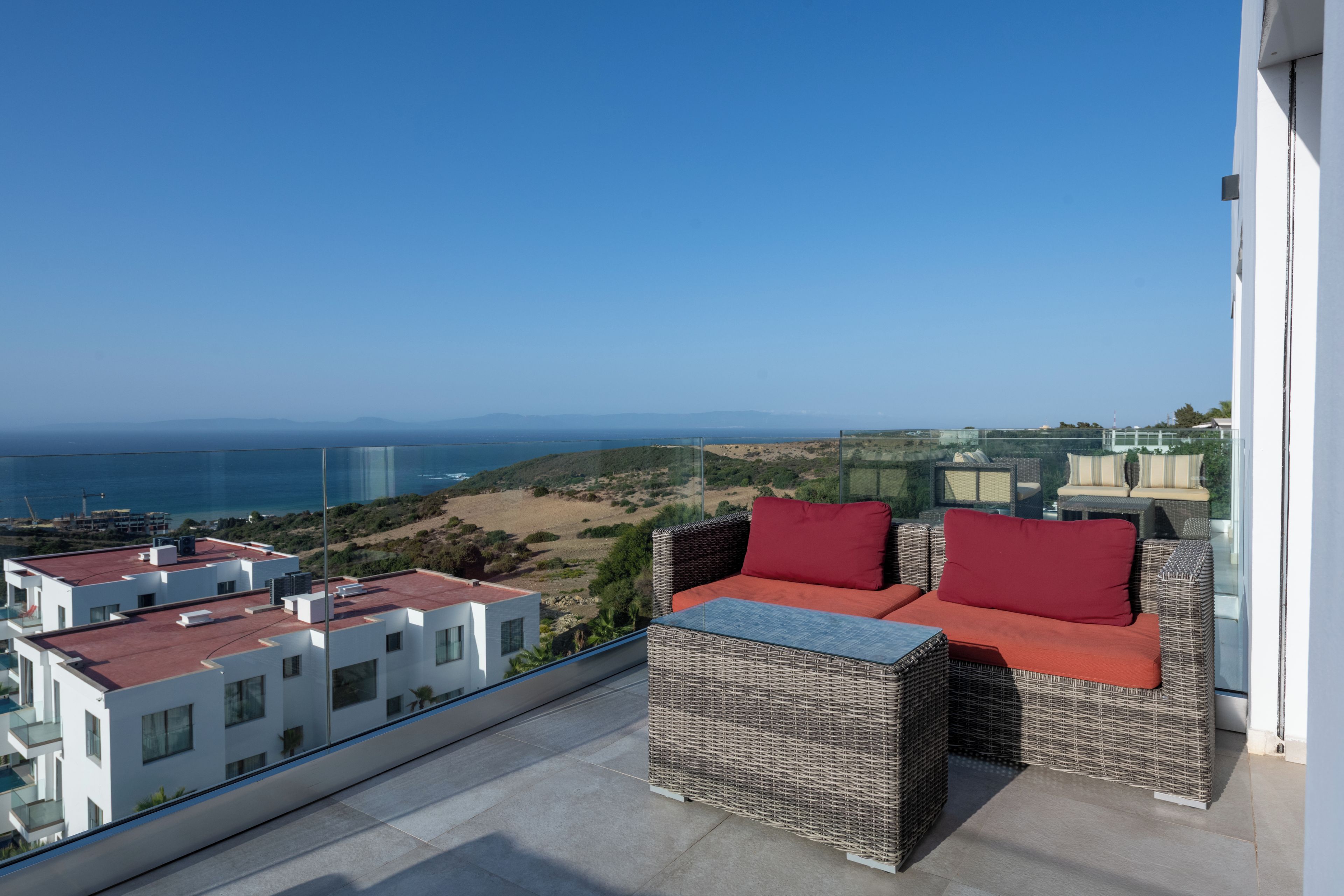 Un balcón con mobiliario de mimbre, vistas despejadas al mar y edificios modernos al fondo.