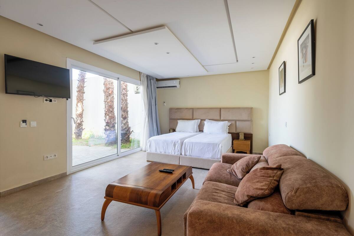 A modern hotel room with a large bed, a sofa, a wooden coffee table, and a TV mounted on the wall.