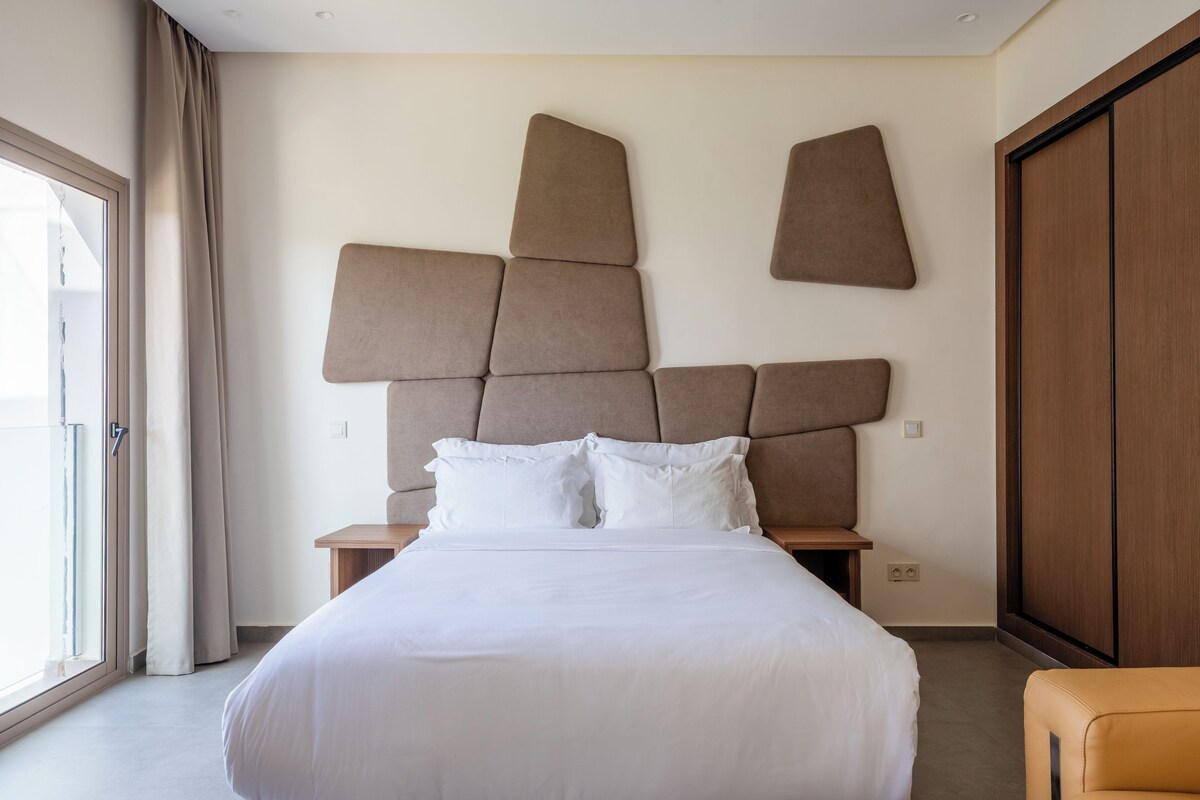 A modern hotel room with a large bed, a headboard with geometric shapes, a wooden nightstand, a wardrobe, and a view through a window with curtains.
