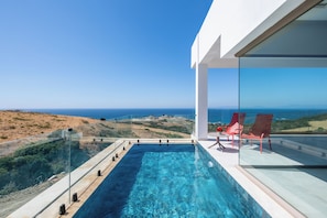 A modern infinity pool with clear blue water, red lounge chairs, and a glass railing overlooking a coastal landscape. - Le Tanger By Mnar Palm (Al Bahraoyine)