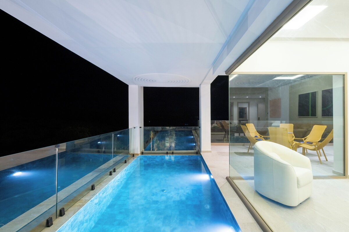 A modern indoor pool area with glass railings, a seating area with yellow chairs, and a view into a dining space.