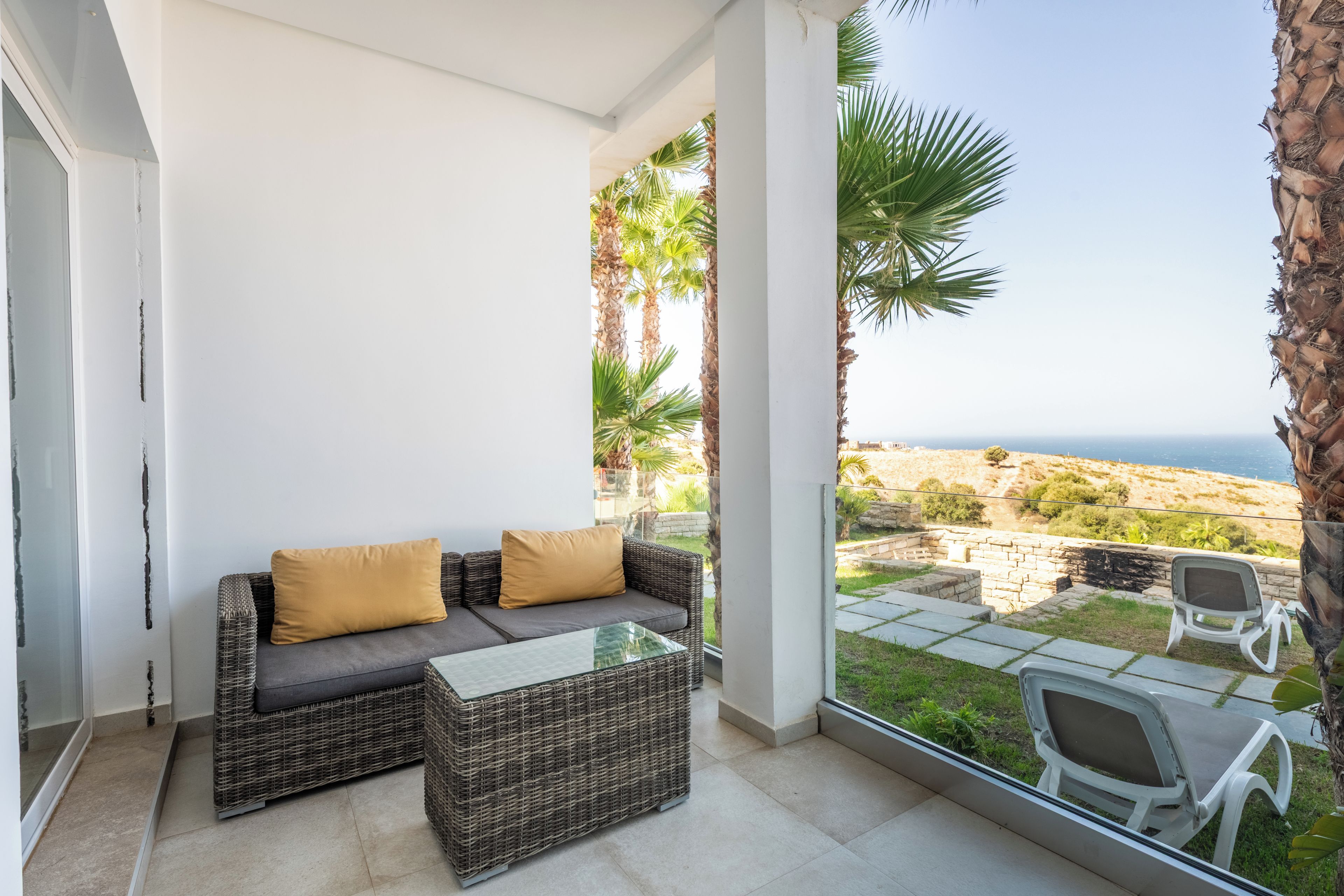A balcony with wicker furniture, a glass table, and a view of the sea.