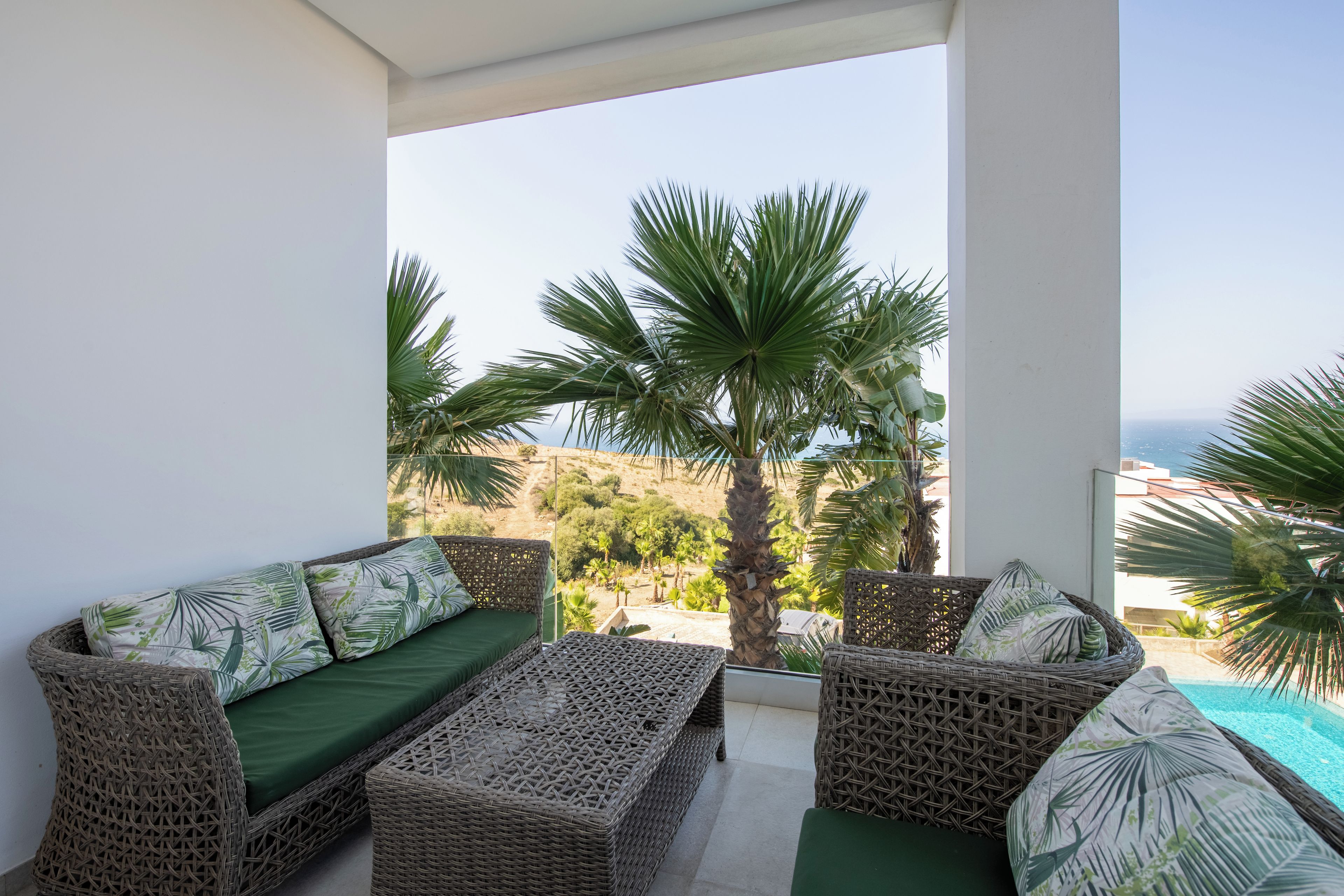 A balcony with wicker furniture, a green cushion, and a view of palm trees and a swimming pool.