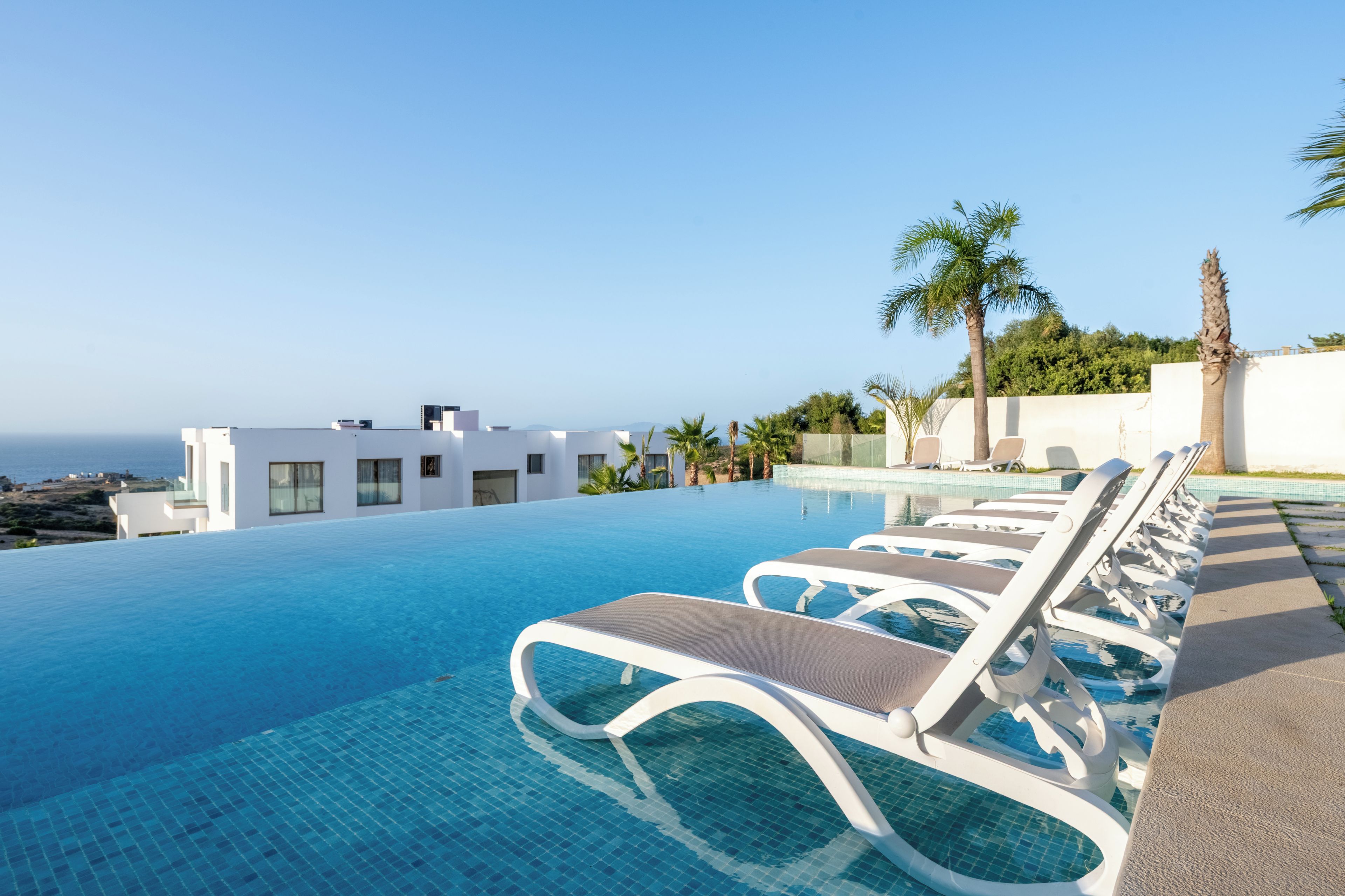 A pool area with lounge chairs, a clear view of the ocean, and a modern building with large windows.